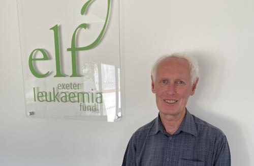 A man smiling stood next to a sign that has the Exeter Leukaemia Fund logo on it